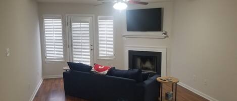A living room with a dark blue sofa, a fireplace, a television mounted above the fireplace, and a ceiling fan.