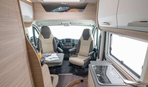 Inside, a central aisle leads to a compact kitchenette with sink and storage, two seating areas with cushioned seats and a steering wheel.