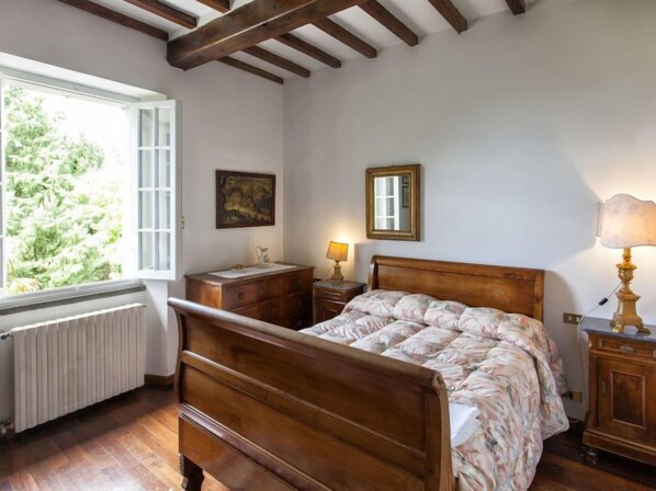 A bedroom with a wooden bed, a nightstand, a dresser, a window with a view of greenery, and a radiator.