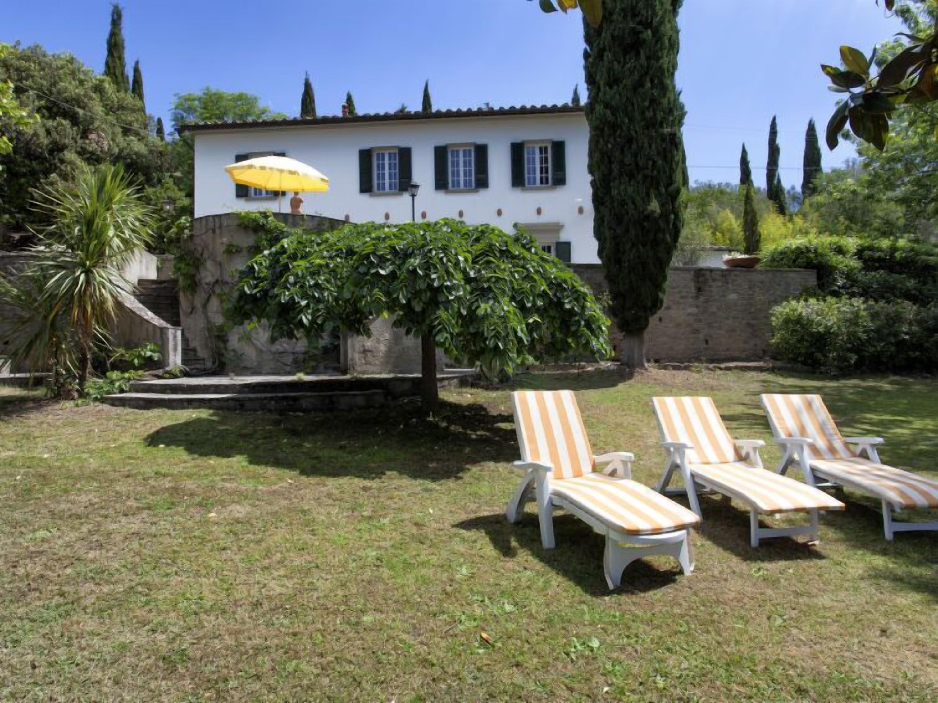A villa with a white exterior, a stone wall, and a large tree in the backyard. There are three striped lounge chairs and a yellow umbrella.