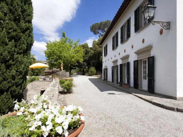 A cobblestone street leading to a white building with black shutters, surrounded by greenery and a statue.