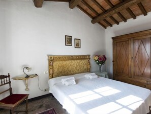 A bedroom with a wooden headboard, a bed with white linens, a wooden wardrobe, a small table with a vase of flowers, and a chair with a red cushion.