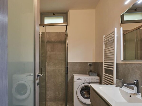 Un baño pequeño con cabina de ducha de vidrio, lavadora y lavamanos con toallero.