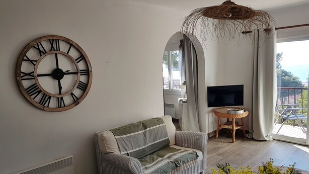 A living room with a large wall clock, a wicker sofa, a TV, and a balcony with a view.