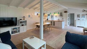 A spacious living area with a high ceiling, a television, a dining table, and a kitchen in the background. - Newly renovated summer house in Hornbæk, close to water and city (Hornbæk)
