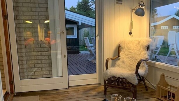 A cozy room with a sliding glass door leading to a wooden deck, a white chair, a small table with a tray, and a lamp. - Newly renovated summer house in Hornbæk, close to water and city (Hornbæk)