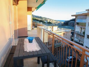 A balcony with a table and a blue trash bin, overlooking a coastal town with buildings and a clear sky.