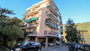 Un immeuble résidentiel à plusieurs étages, doté de balcons, avec un espace de stationnement à l’avant et une colline en arrière-plan.