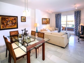 A modern living room with a glass dining table, a beige sofa, and a large window with curtains.