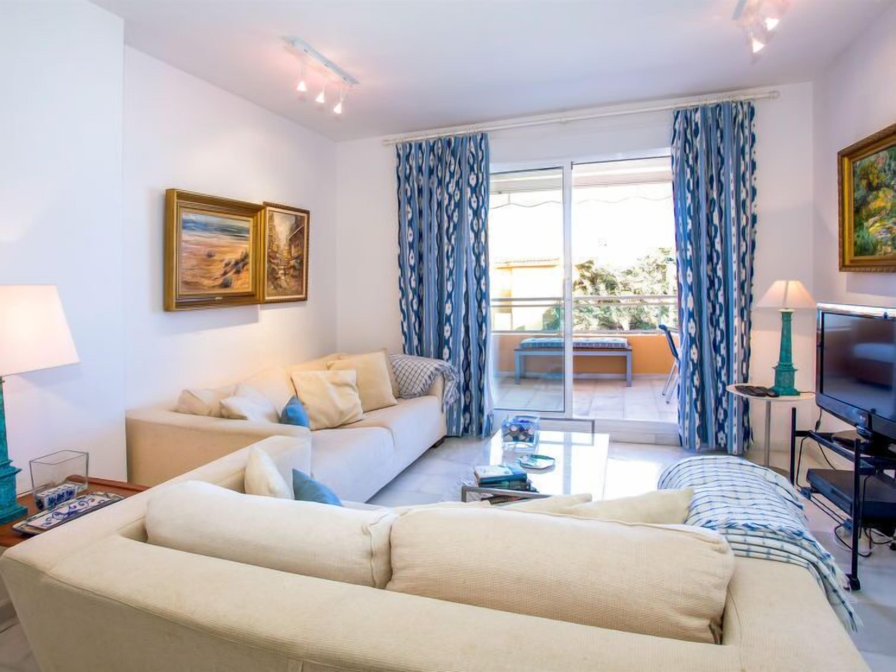 Ein Wohnzimmer mit Sofa, Couchtisch und Fernseher. Vorhänge und ein Gemälde sind an der Wand zu sehen.