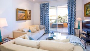 A living room with a sofa, a coffee table, and a TV. There are curtains and a painting on the wall.