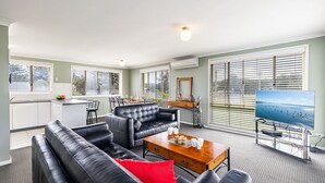 Ett modernt vardagsrum med en svart lädersoffa, ett träbord och en tv. Det finns också en köksdel med ett matbord och stolar. Stora fönster med persienner ger naturligt ljus och fin utsikt.