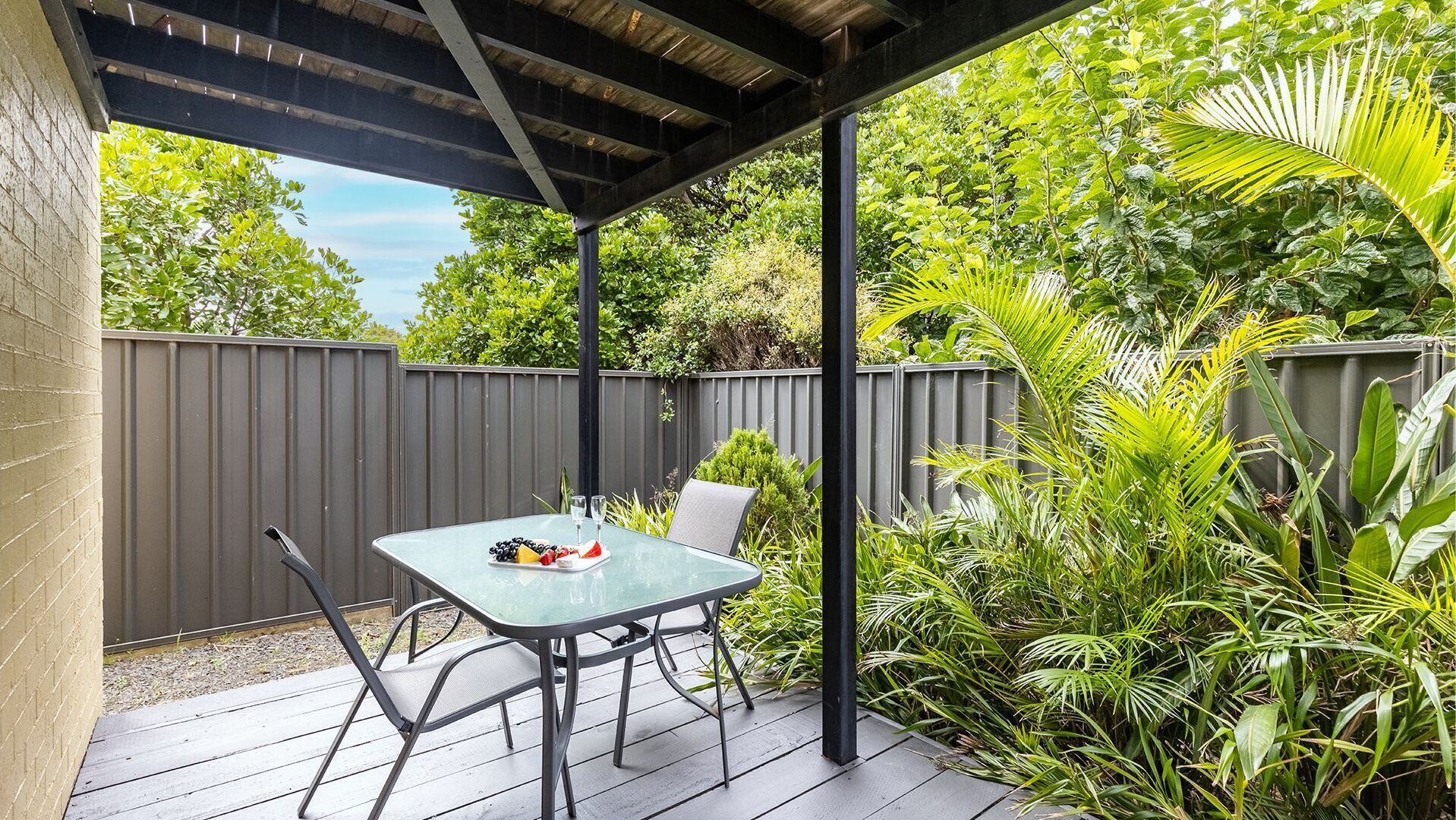 Un patio exterior con una mesa lista para dos personas, rodeado de abundante vegetación y una terraza de madera.