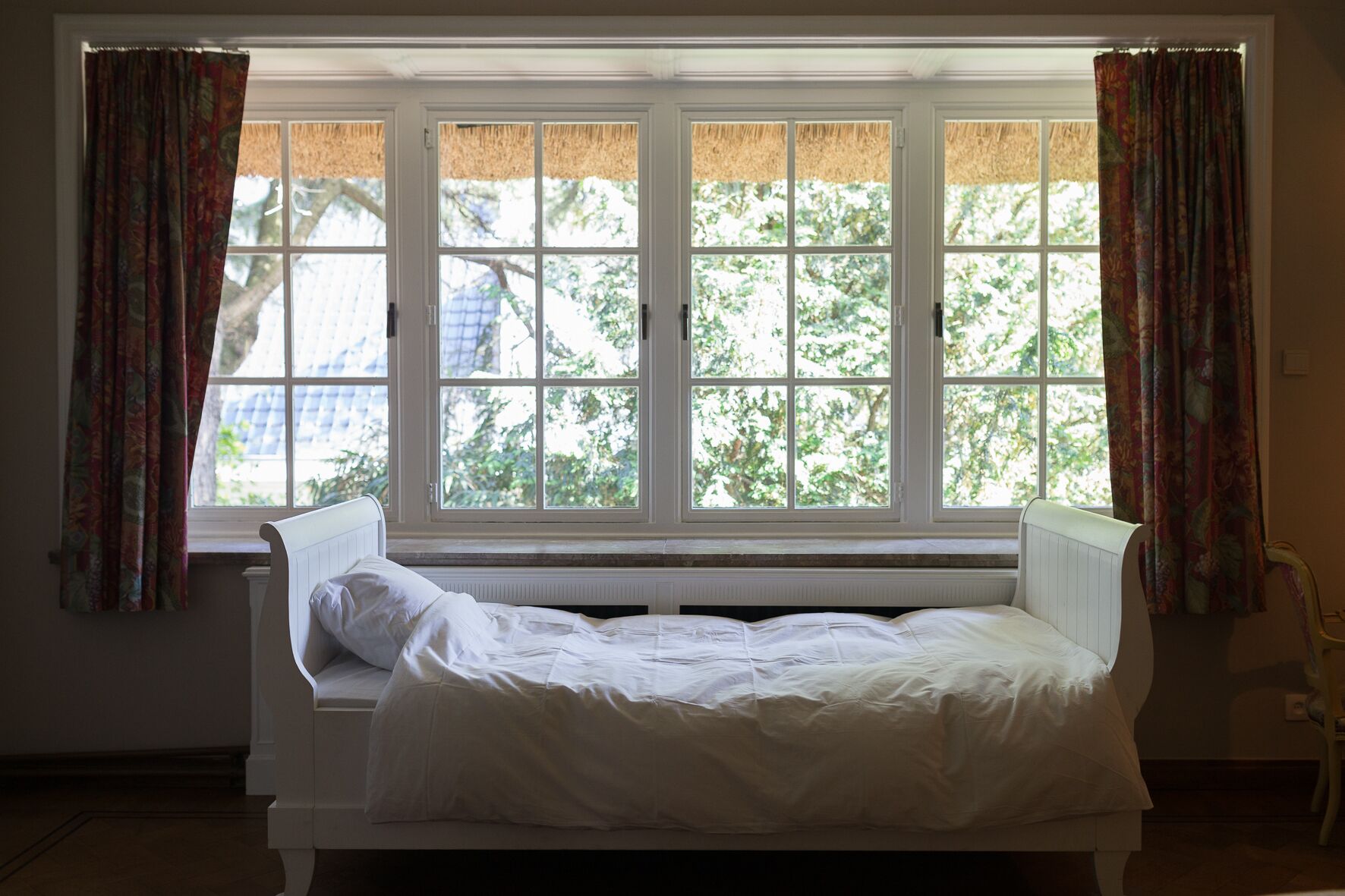 A single bed with a white headboard and footboard, positioned under a large window with a view of trees.