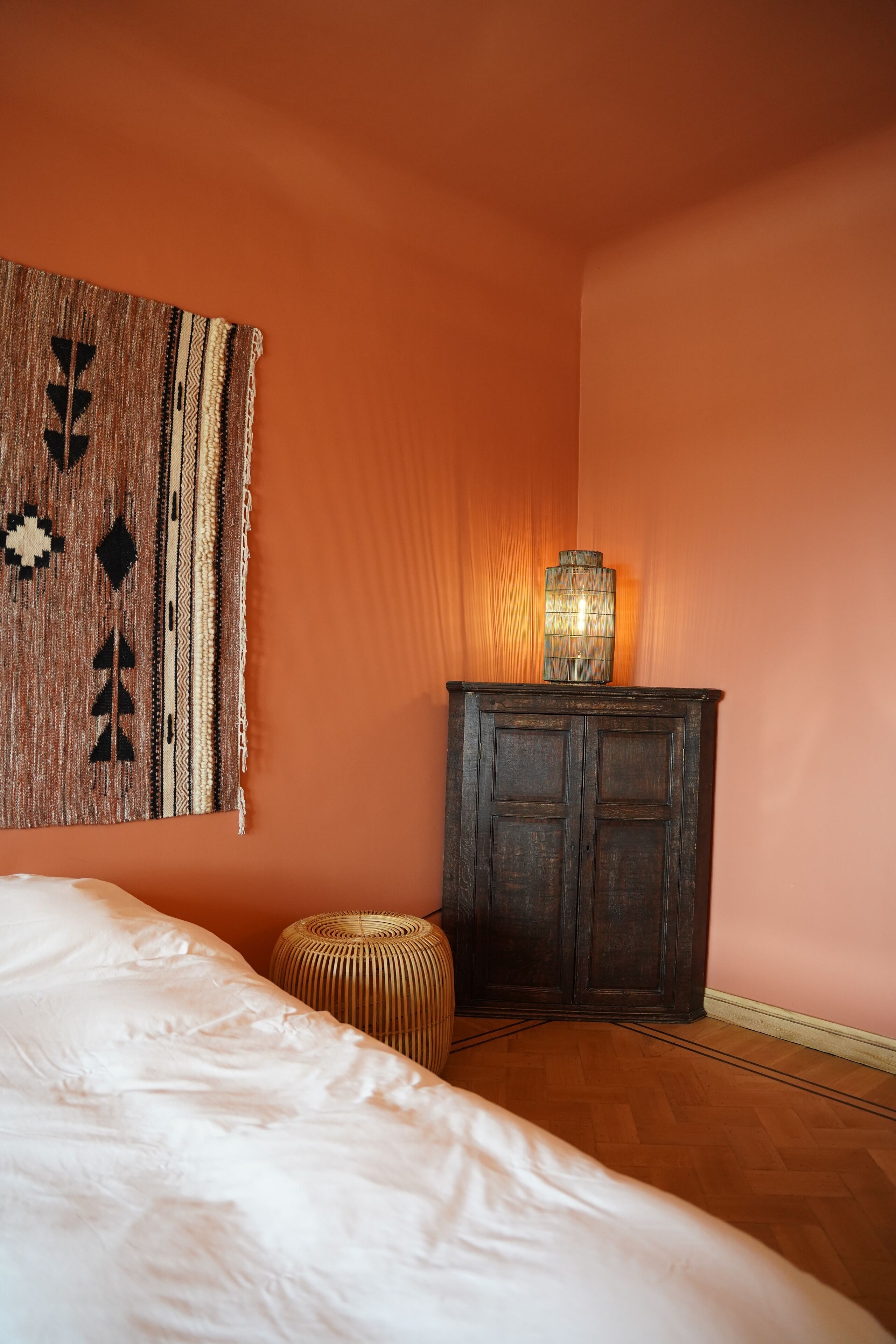 A bedroom with a bed, a wooden cabinet, a woven stool, and a patterned wall hanging.