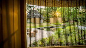 Blick durch ein Fenster mit vertikalen Jalousien, der ein Gebäude mit Strohdach, einen kleinen Teich und umgebende Grünflächen zeigt.