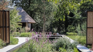 Ein ruhiger Garten mit einem strohgedeckten Gebäude, einem schmalen Wasserlauf und üppigem Grün.