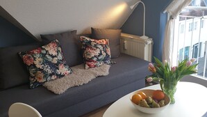 Una habitación acogedora con un sofá gris, cojines estampados, una mesa redonda blanca y un frutero, y vistas a edificios a través de una ventana.