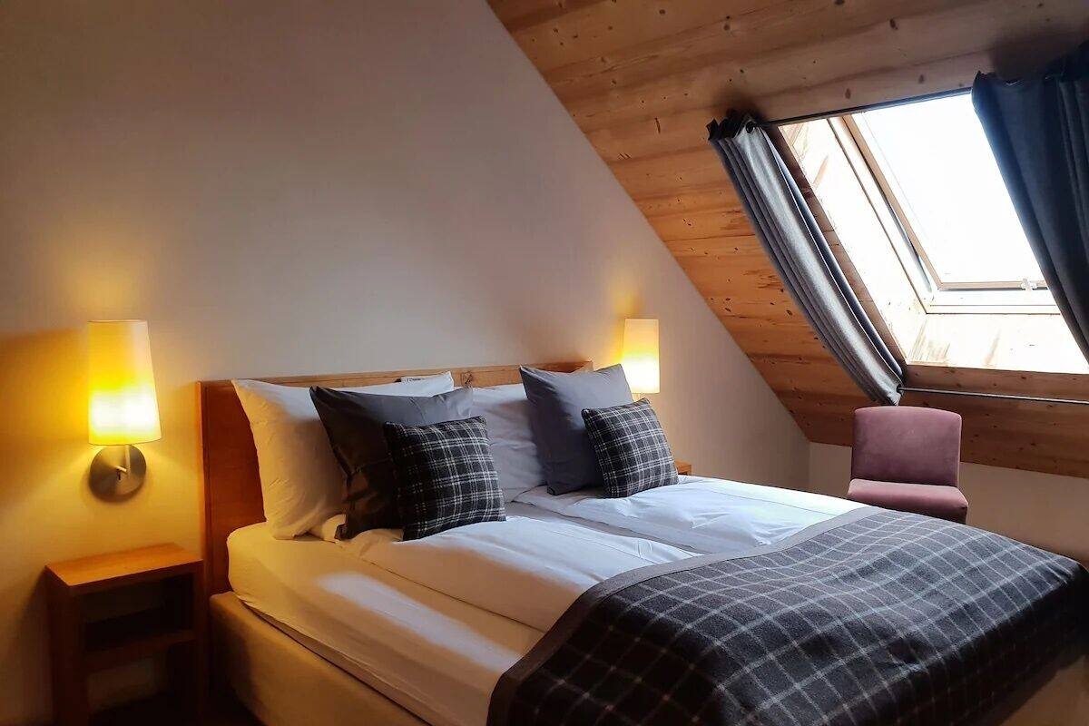 Ein gemütliches Schlafzimmer mit schrägem Dach, einem Bett mit weißer Bettwäsche, zwei Kissen, einem Nachttisch mit Lampe und einem Fenster mit Vorhängen.