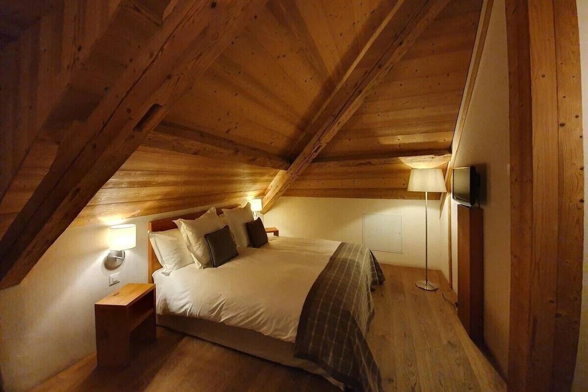 Ein gemütliches Schlafzimmer im Dachgeschoss mit Holzbalkendecke, einem Bett mit Kissen, einem Nachttisch, einer Lampe und einem Fernseher.
