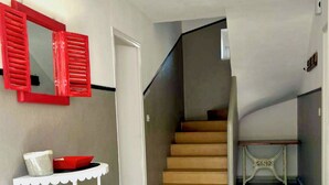 Un escalier menant à l’étage supérieur, une console blanche sur laquelle repose un bol rouge, et un miroir encadré de rouge accroché au mur.