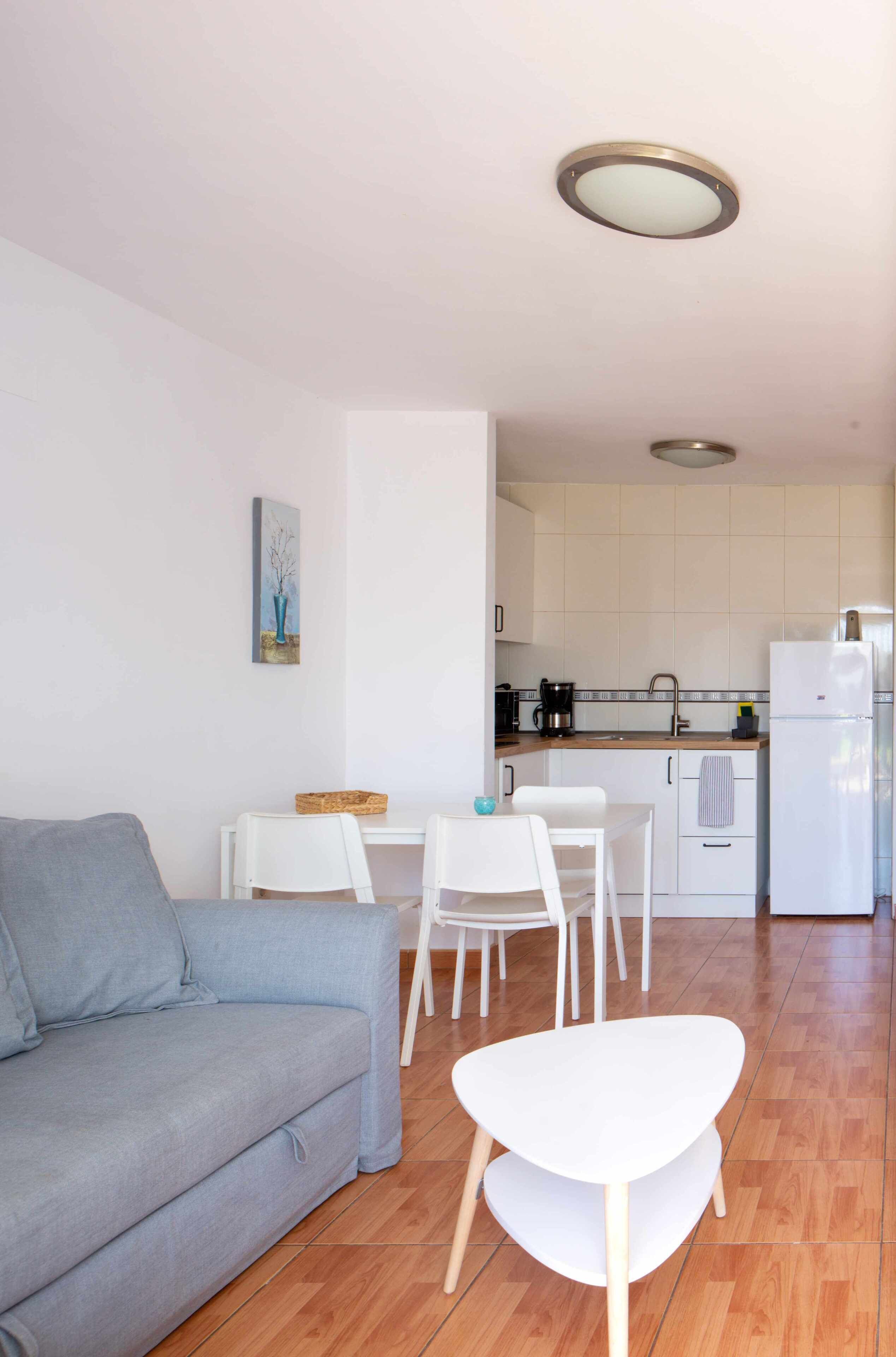A modern living room with a grey sofa, a white table, and a white chair. A kitchen with white cabinets and a white refrigerator is visible in the background.