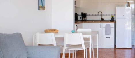 A modern living room with a grey sofa, a white table, and a white chair. A kitchen with white cabinets and a white refrigerator is visible in the background.