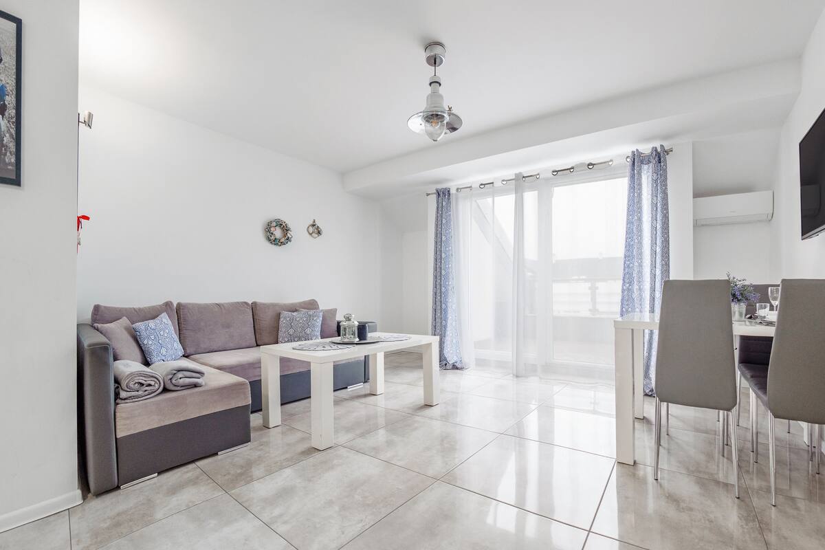 Una sala de estar moderna con sofá, una mesa de comedor y sillas. Hay un televisor montado en la pared y un ventilador de techo.