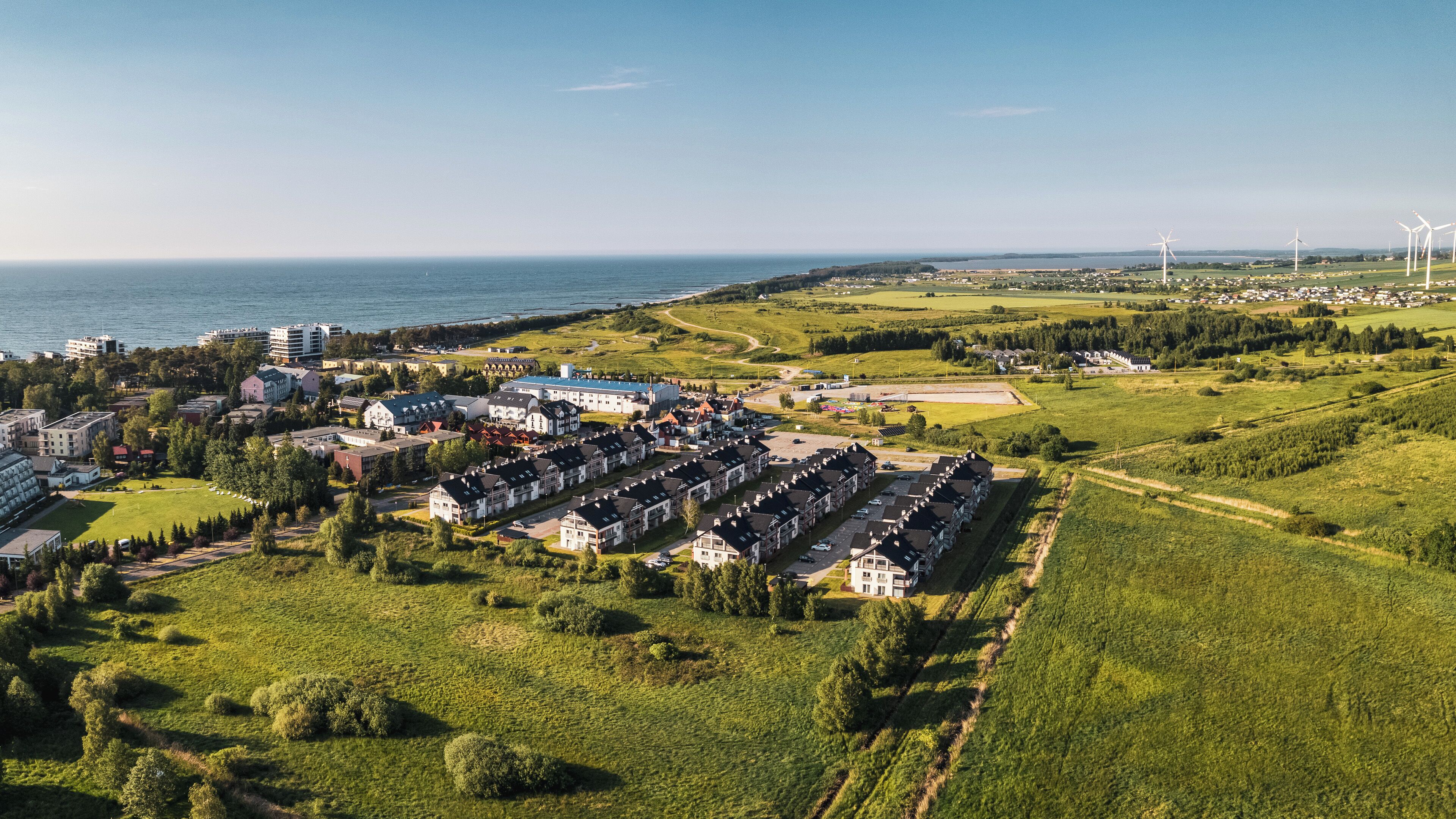 An aerial view of a coastal town with residential buildings, a marina, and wind turbines in the distance.