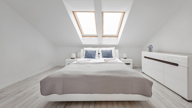 Un dormitorio moderno con tragaluz, una cama con ropa de cama blanca, una mesita de noche con lámpara y un armario blanco.