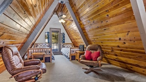 Um quarto de sótão aconchegante, com ripas de madeira no teto, duas poltronas de couro, um banco de madeira e vista para o exterior através de uma porta.