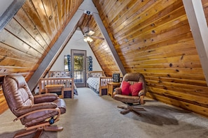 A cozy attic bedroom with wooden slats on the ceiling, a ceiling fan, a bed with a polka dot bedspread, and two leather chairs.