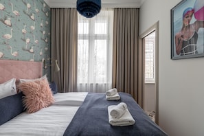 Ein Hotelzimmer mit Bett, einem großen Fenster mit Vorhängen, einem Bild an der Wand und einer blauen Pendelleuchte.