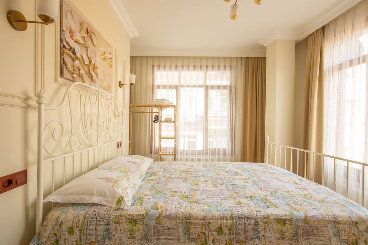 A neatly made bed with a patterned bedspread, a wall-mounted shelf with a towel, and a large window with curtains.