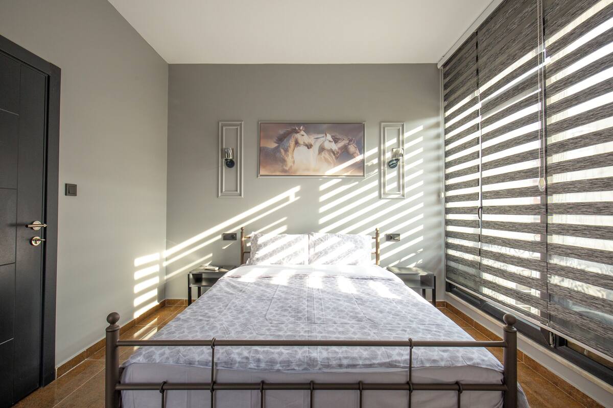 A bedroom with a bed, a painting on the wall, and a window with blinds.