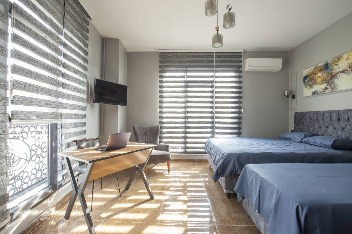 A bedroom with two beds, a desk with a laptop, a TV on the wall, and a window with blinds.