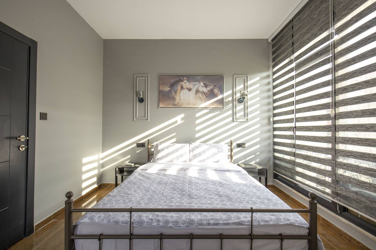 A bedroom with a bed, a painting on the wall, and a window with blinds.