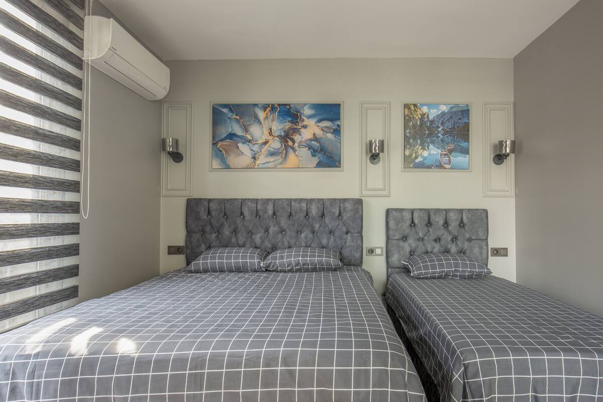 A bedroom with two beds, a large window with blinds, and two wall-mounted paintings.
