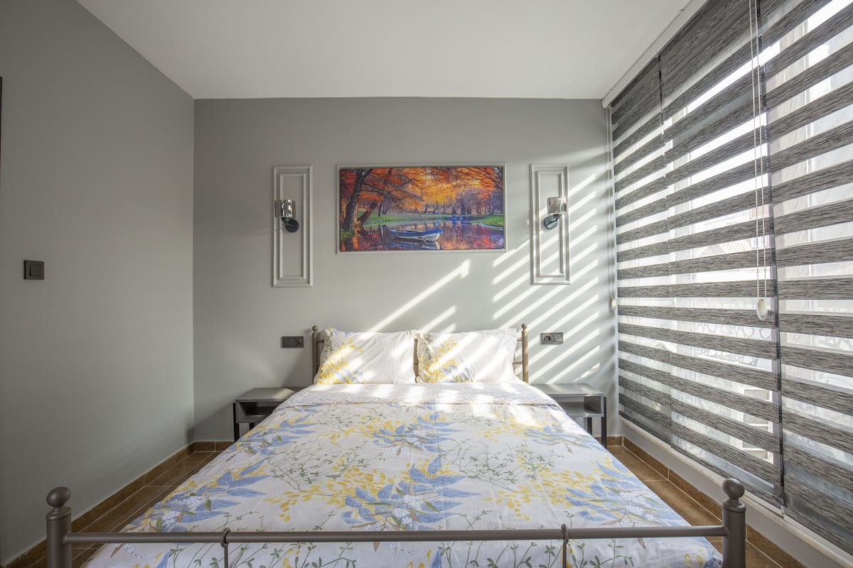 A bedroom with a bed, a painting on the wall, and a window with blinds.