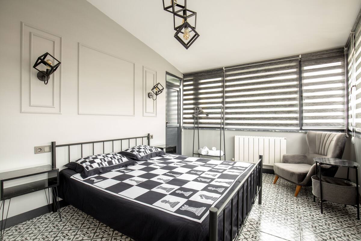 A modern bedroom with a checkered bedspread, a grey armchair, a small table, and a radiator.