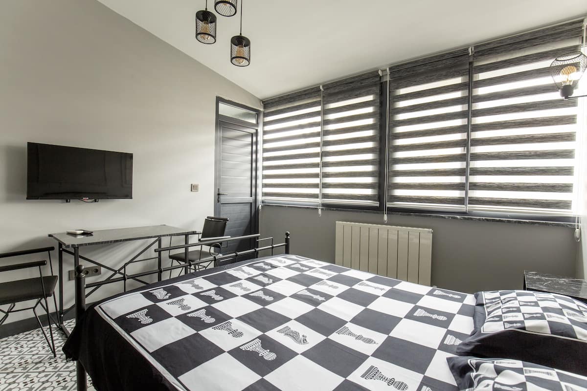 A bedroom with a checkered bedspread, a desk with a chair, a television on the wall, and large windows with blinds.