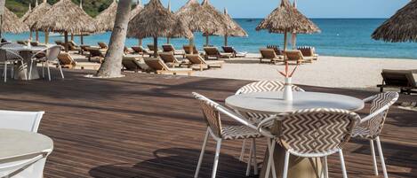 A beachside restaurant with outdoor seating, including tables and chairs, and thatched umbrellas.