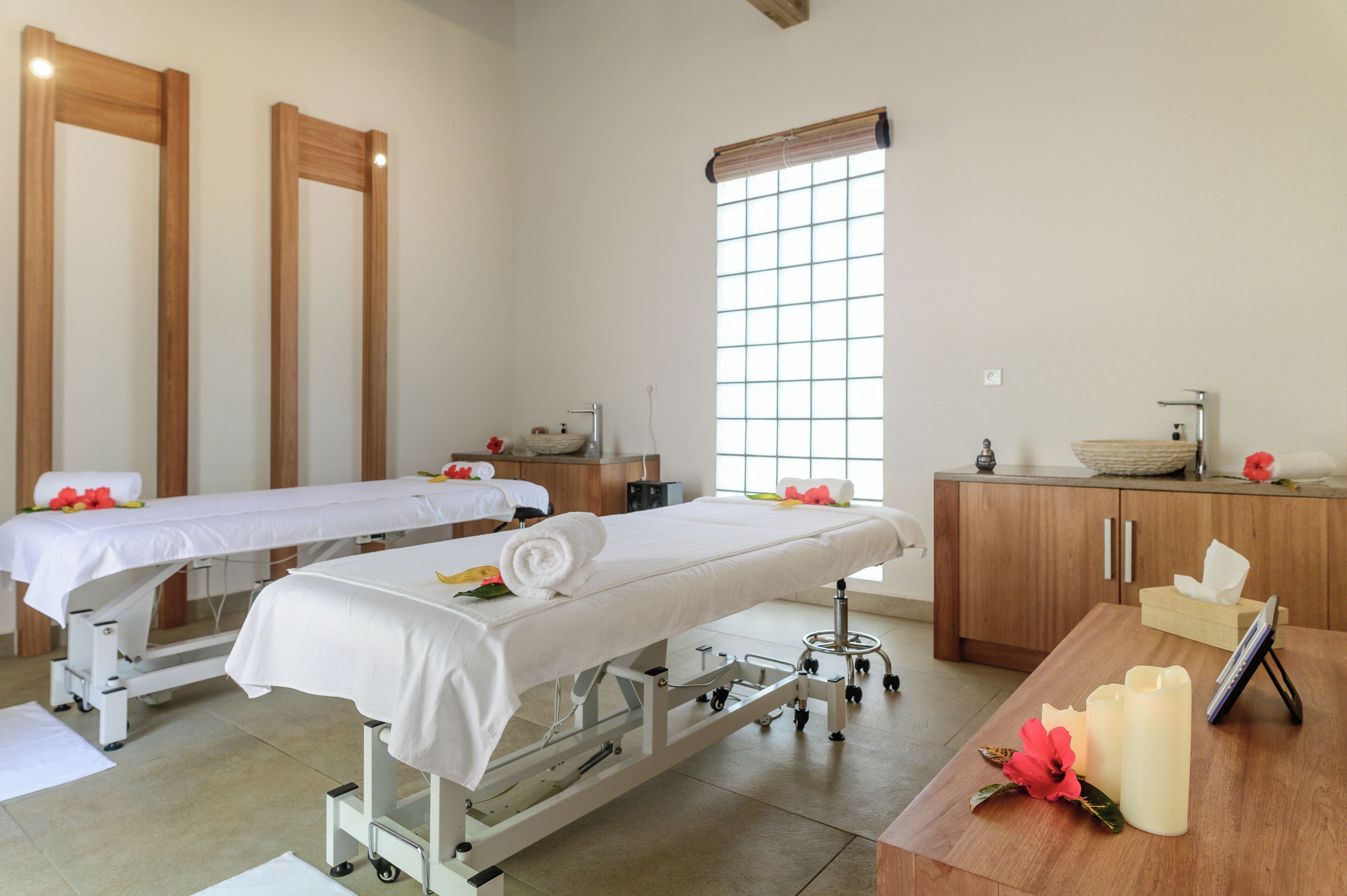 A spa room with two treatment beds, a wooden cabinet, a window, and a table with candles and flowers.