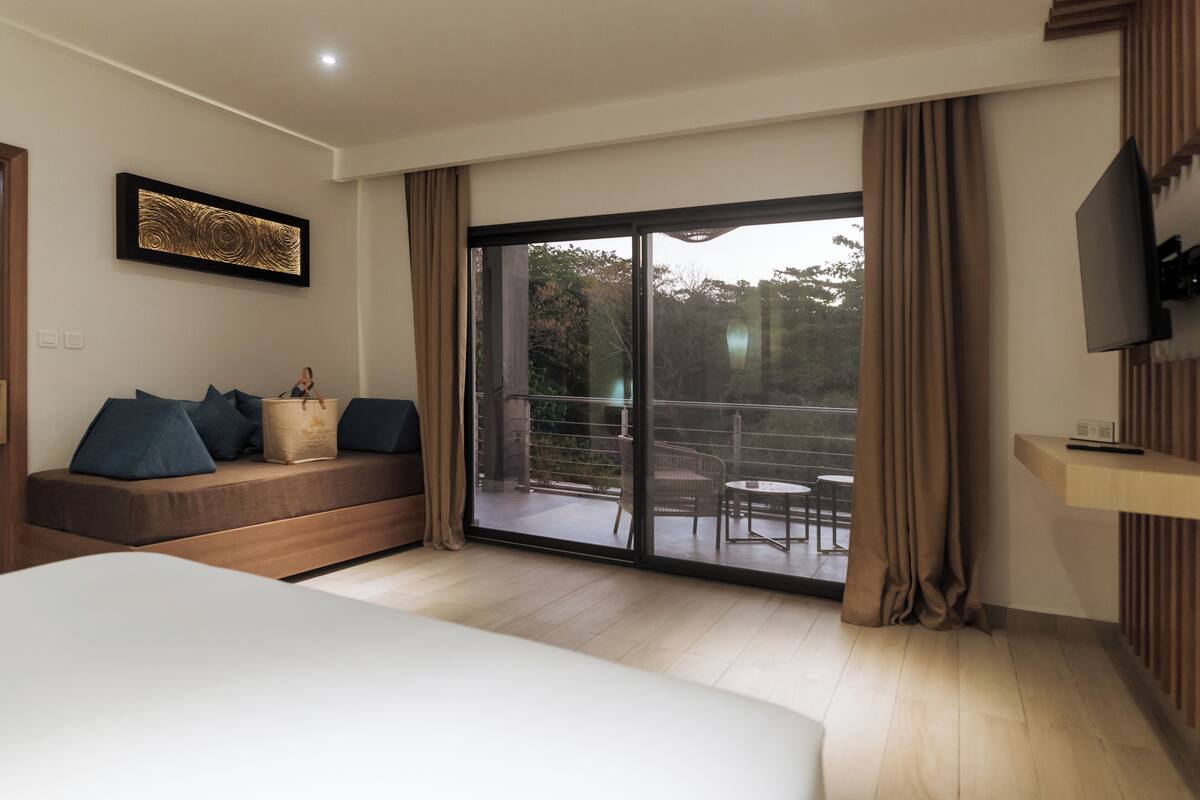 Una habitación de hotel con una cama grande, un sofá con cojines, un televisor y un ventanal corredero que da a un espacio exterior con una mesa y sillas.