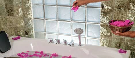 Una bañera llena de agua y pétalos de flores rosados, con una mano agregando más pétalos desde una cesta.