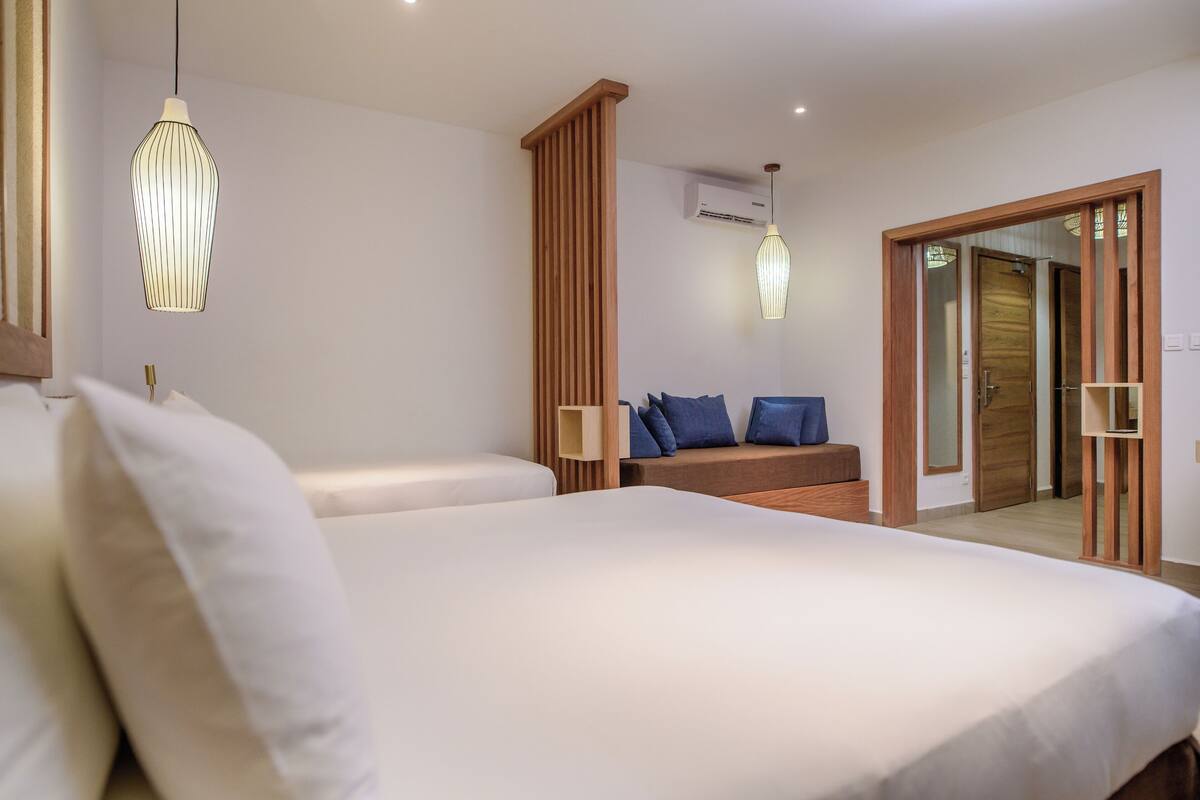 Un dormitorio moderno con una cama grande, un banco de madera con cojines, una lámpara colgante y una puerta que da a otra habitación.