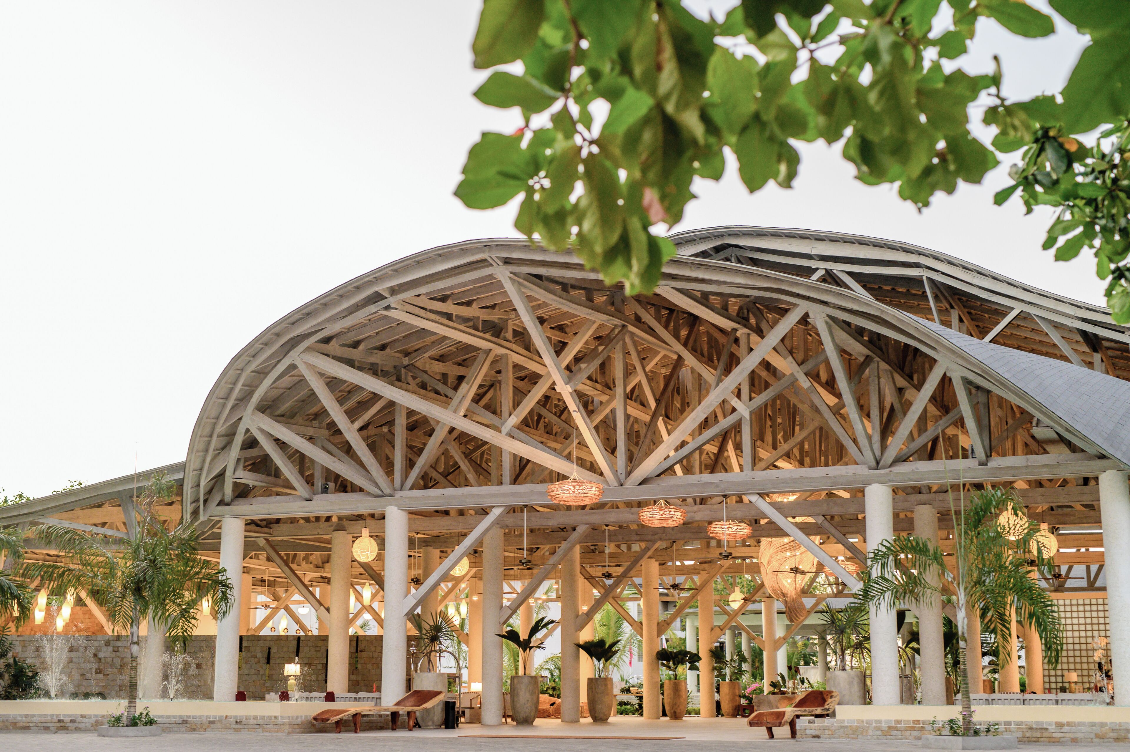 A large, open-air structure with a wooden framework and columns, surrounded by potted plants and outdoor seating areas.