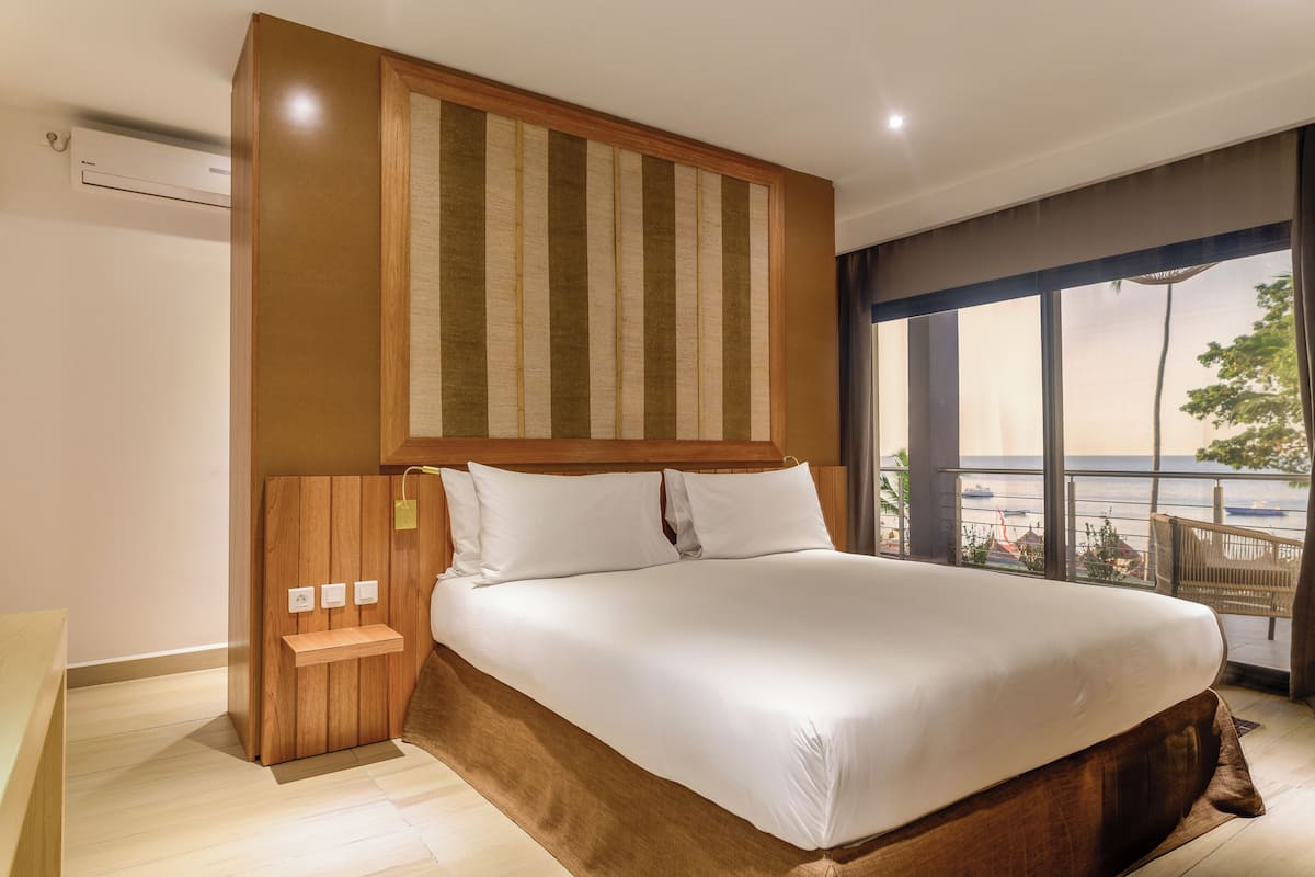 Una habitación de hotel moderna con una cama grande, un cabecero de madera, una mesita de noche pequeña y vistas al mar a través de amplios ventanales.
