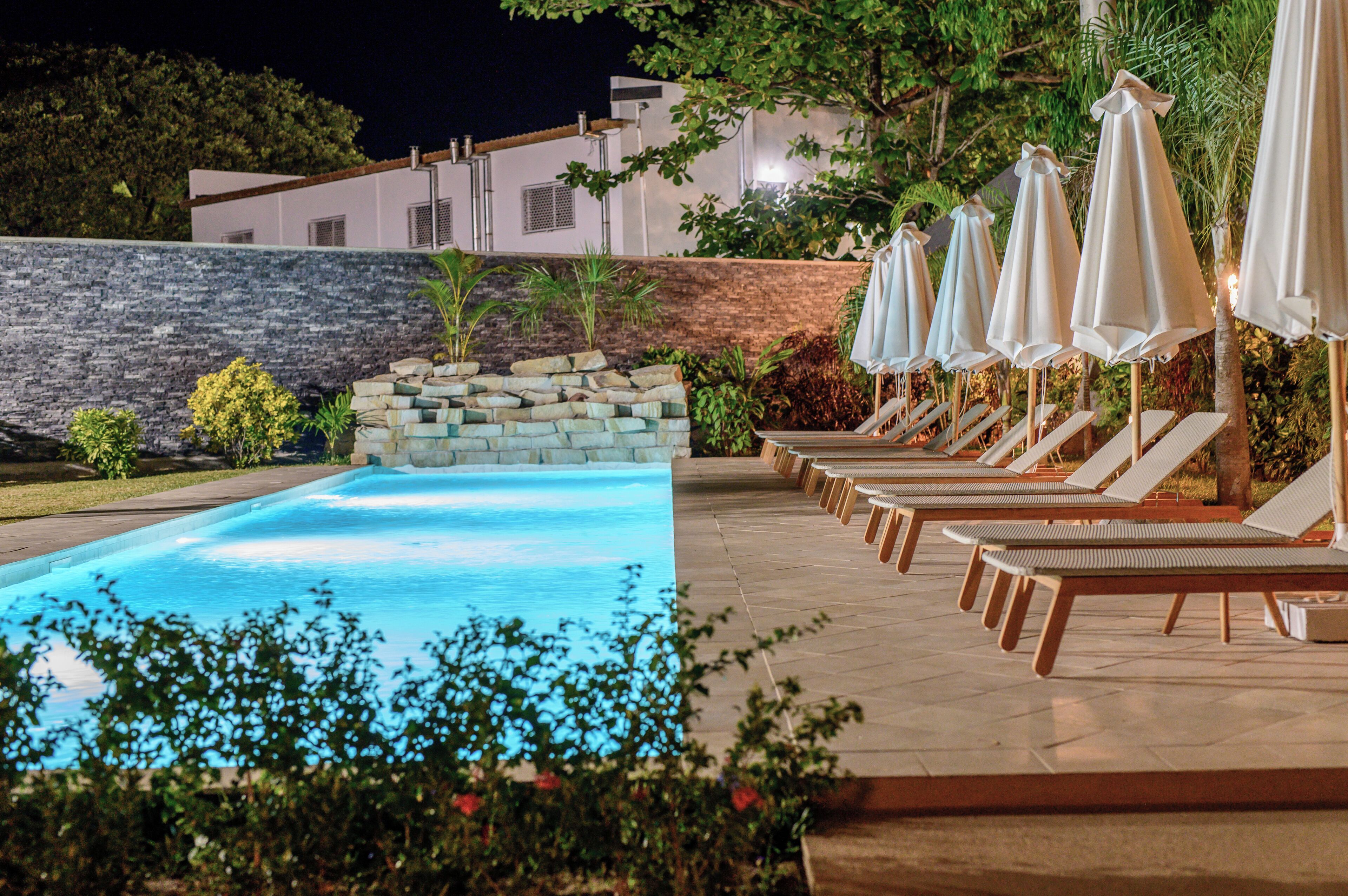 A swimming pool with lounge chairs and umbrellas, surrounded by a stone wall and greenery.