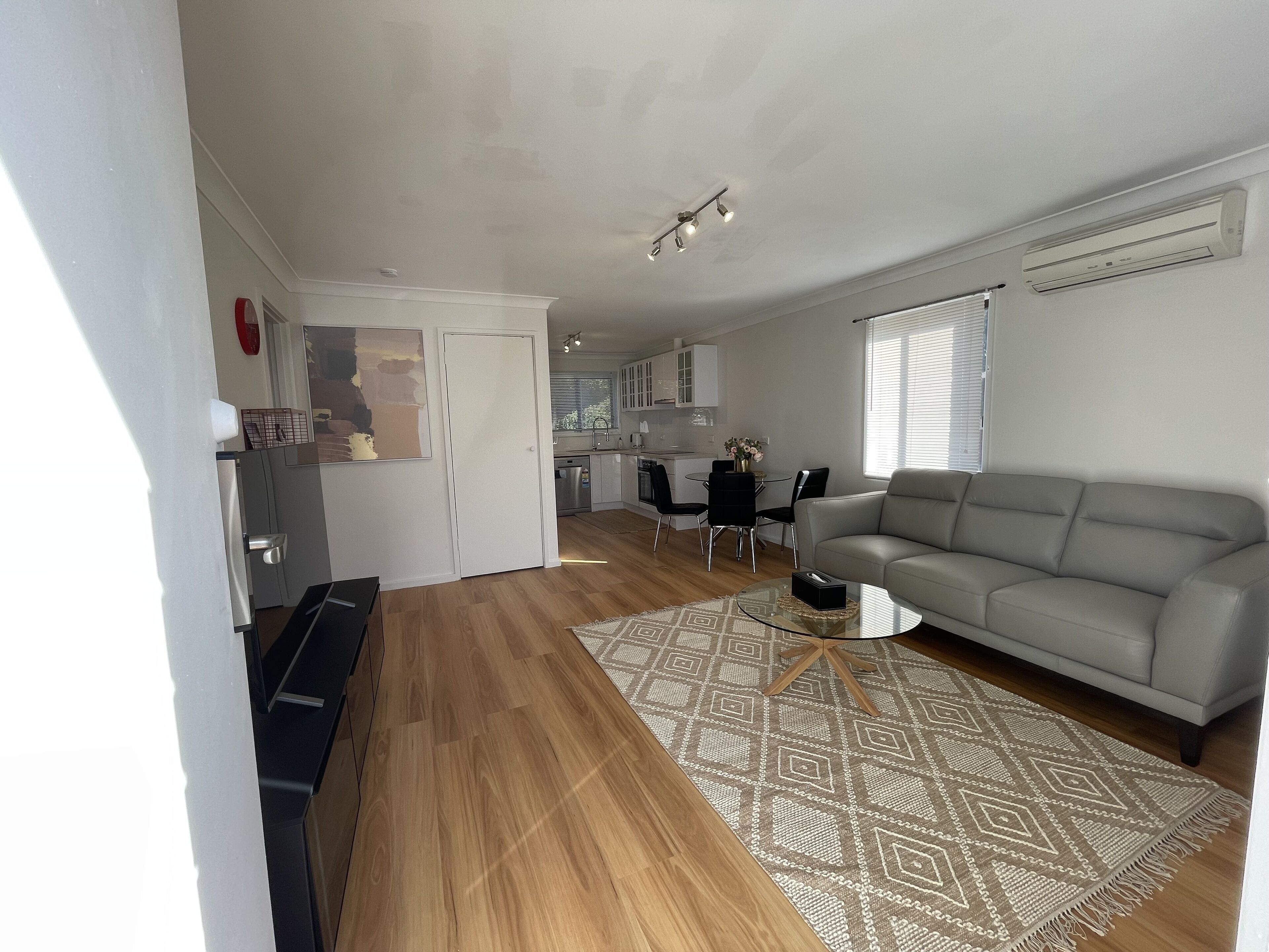 Una sala moderna con un sofá gris, una mesa de centro y un tapete estampado. Se ve una cocina con armarios blancos y una zona de comedor con una mesa y sillas al fondo.